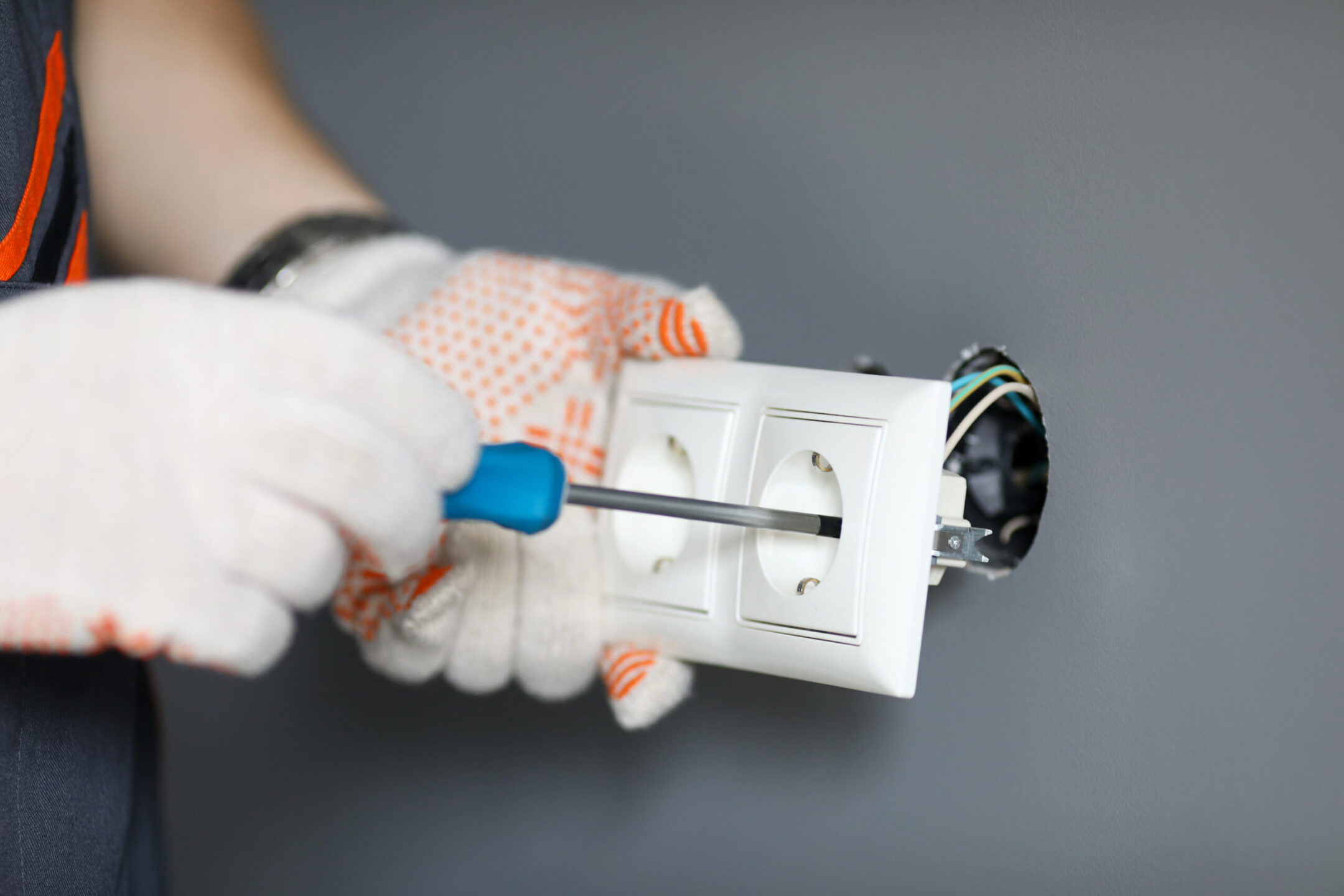 Repairman in gloves fixing socket with screwdriver over grey wall background.